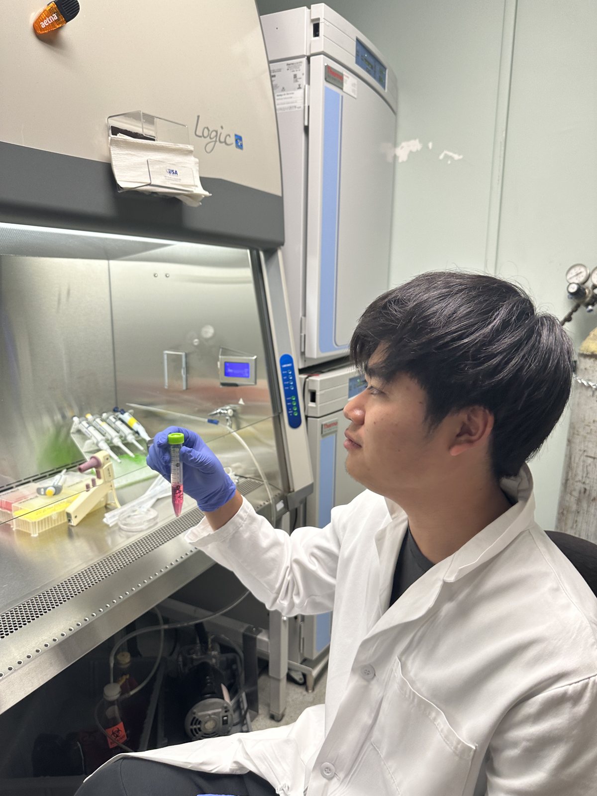 Jalen in a white lab coat and gloves working at a biosafety cabinet, holding a sample tube up to the light