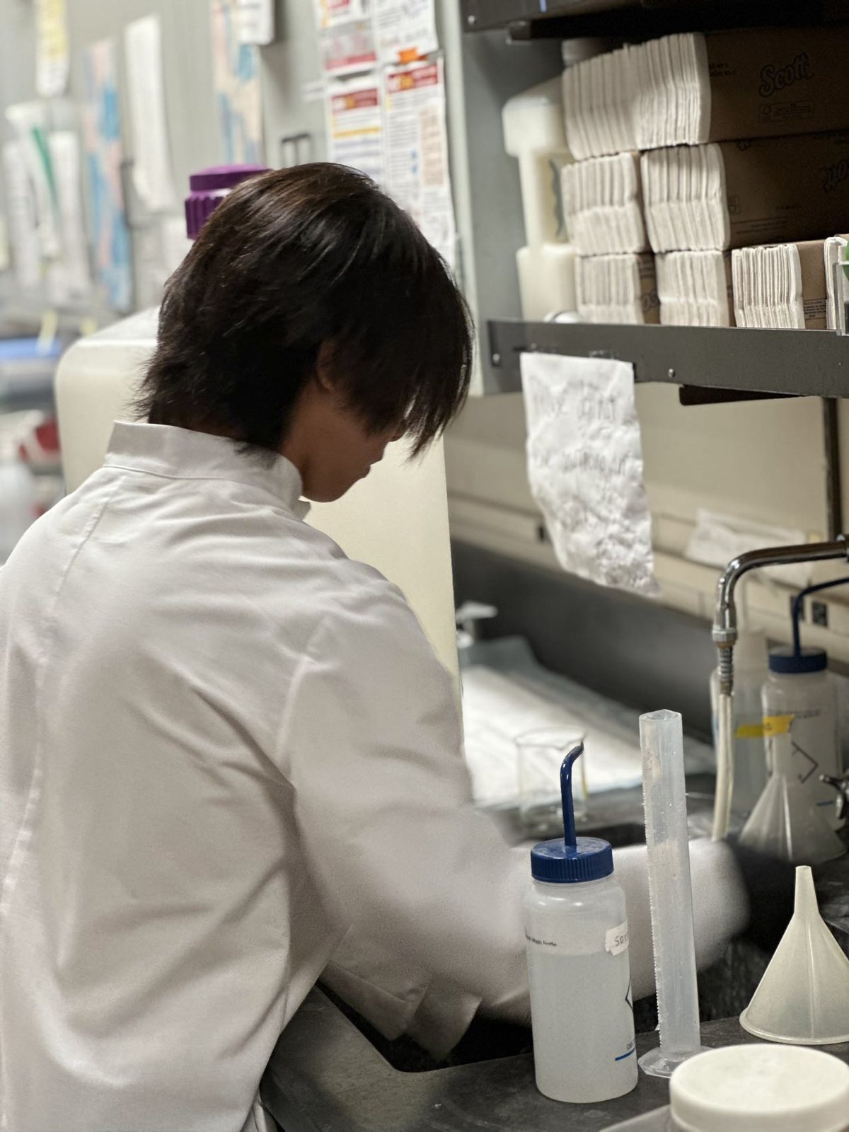 Jalen in a white lab coat at a lab sink, a funnel and a wash bottle on the counter