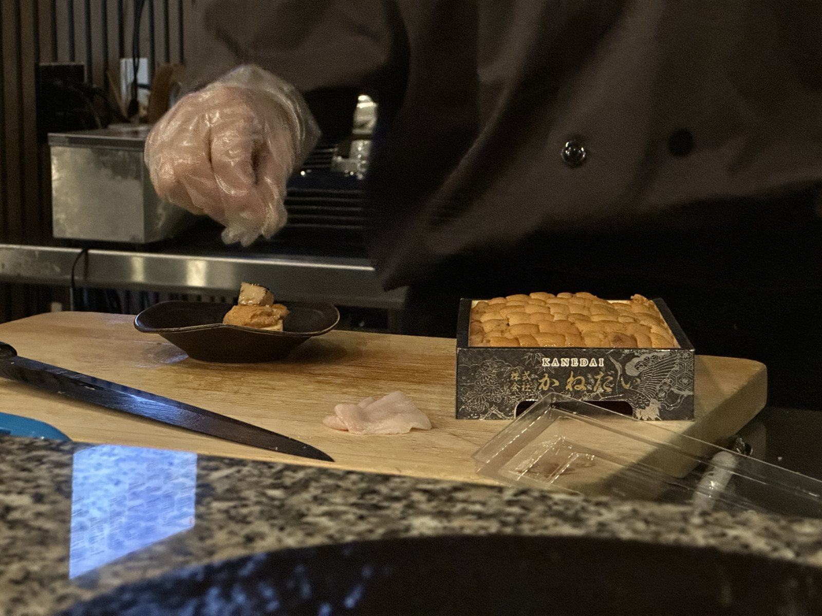 A chef's gloved hands at a sushi counter, a wooden box of nigiri waiting on the board
