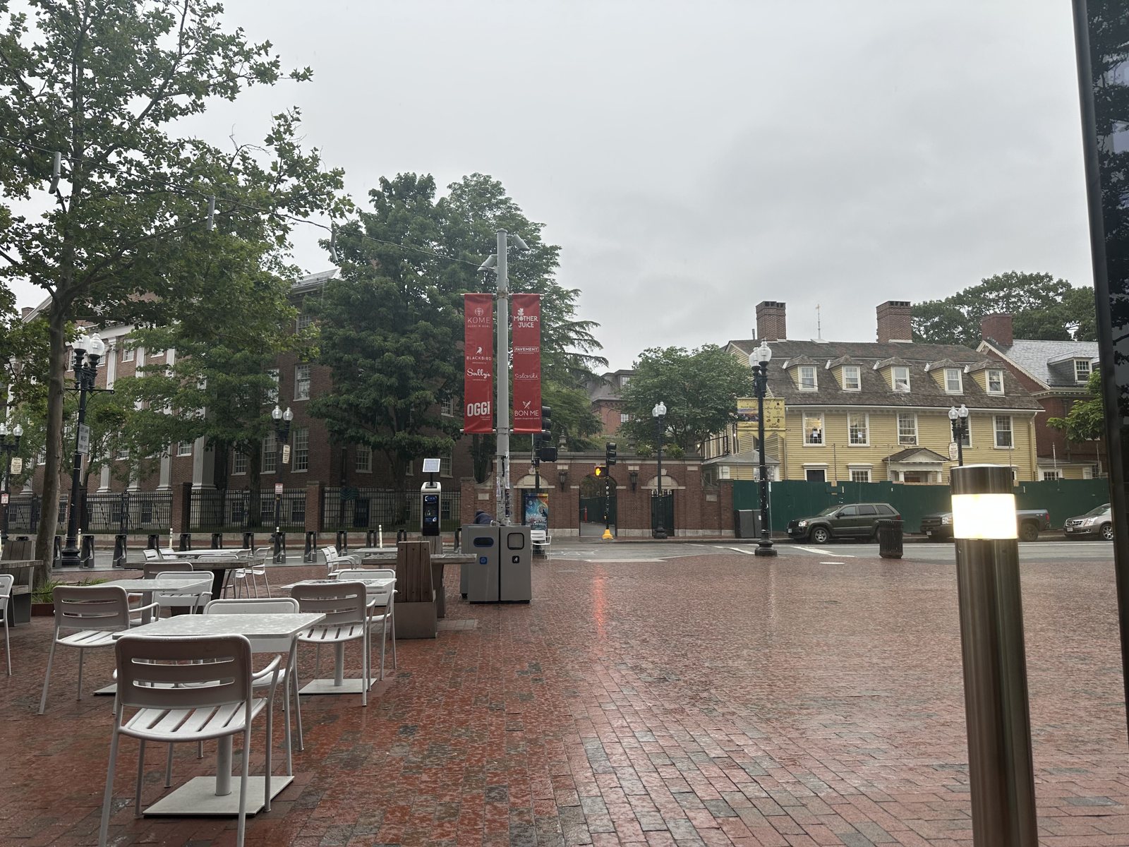 A rain-slicked brick plaza with empty cafe tables, crimson Harvard banners on the lampposts