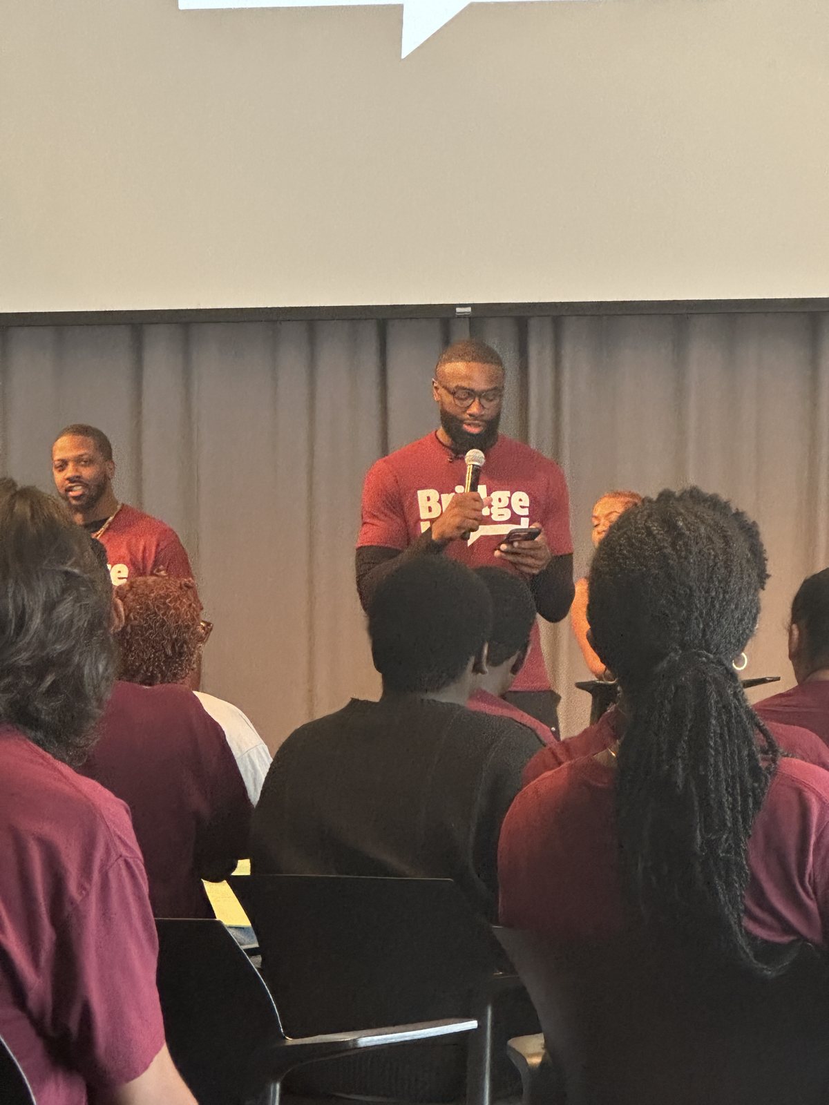 Two speakers in maroon Bridge T-shirts addressing a room of students, seen from the audience