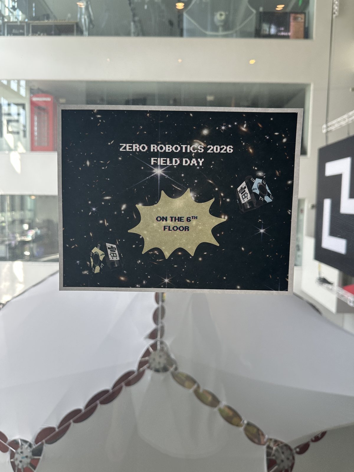 A printed sign reading Zero Robotics 2026 Field Day, on the 6th floor, with two Astrobee robots drawn on a starfield