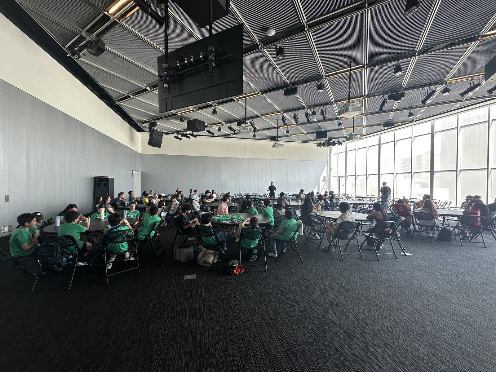 A high-ceilinged room at MIT on Zero Robotics field day, students in green T-shirts at round tables, a wall of windows at the far end