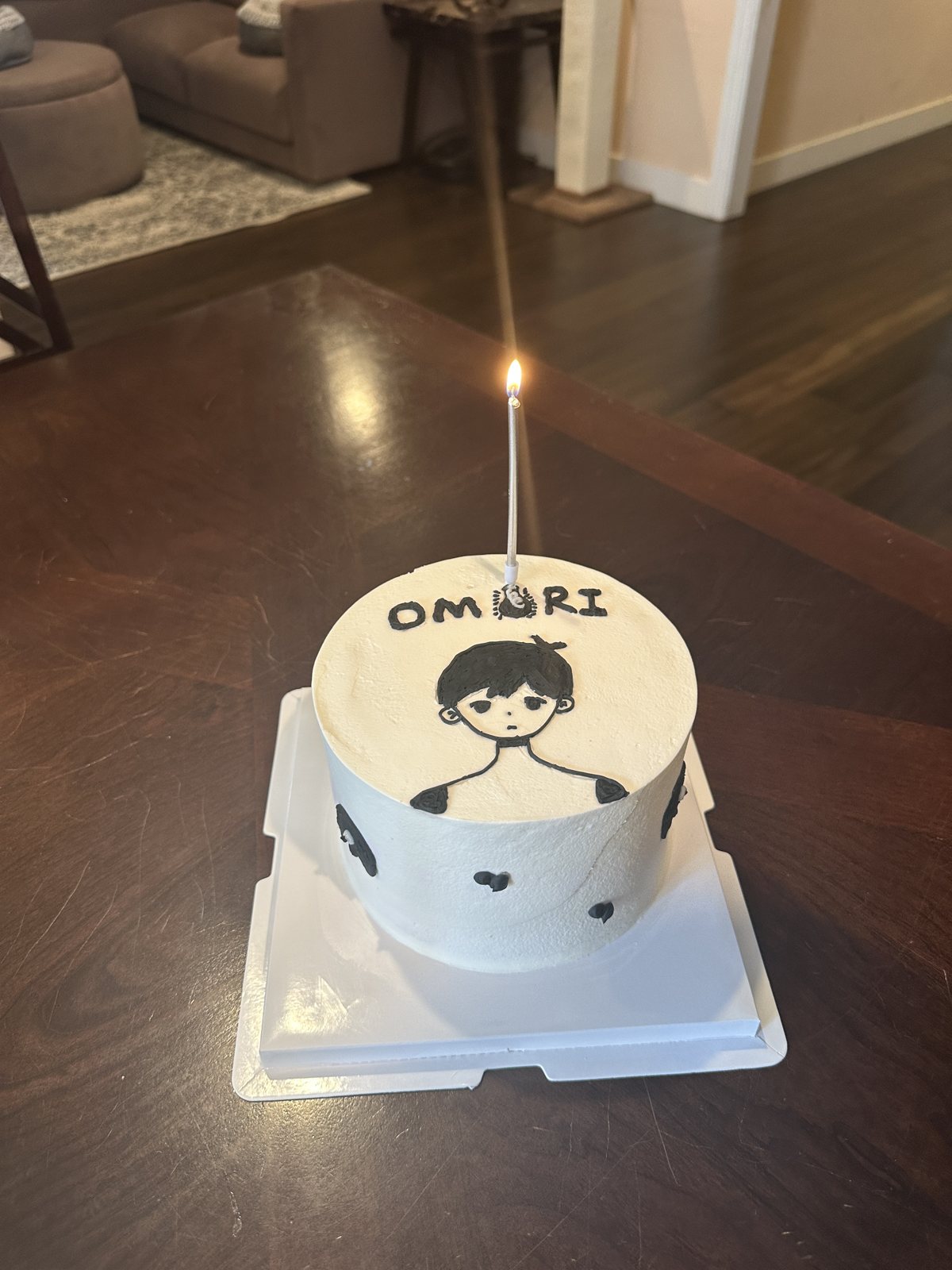 A small white birthday cake with a single lit candle, decorated with a drawing of Omori and the word OMORI