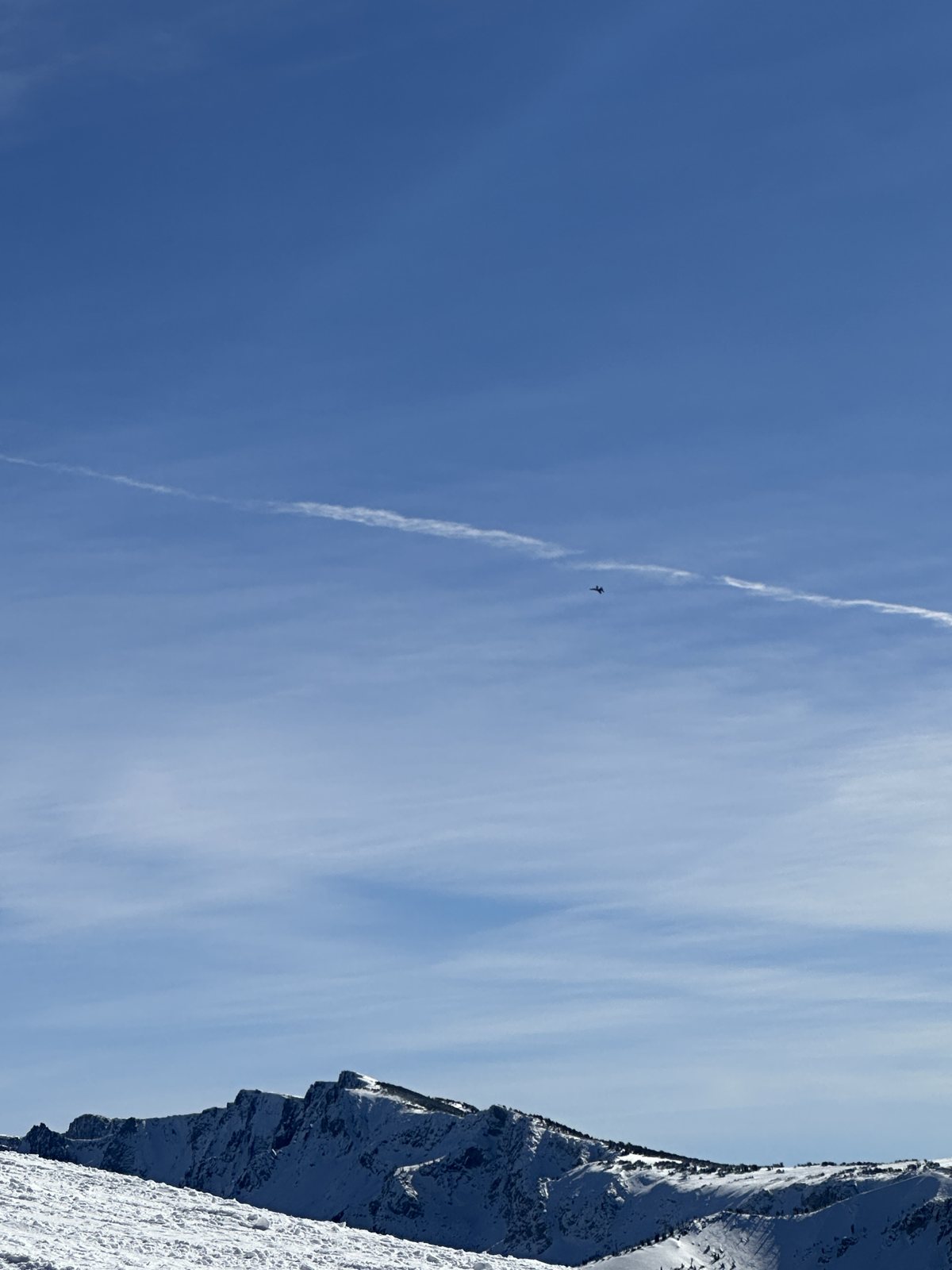 A snow-covered ridge line under a deep blue sky, a single contrail overhead