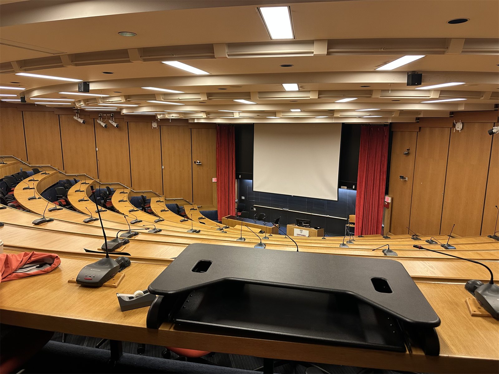 An empty tiered lecture hall at Harvard with wood-panelled desks, a projection screen and red curtains at the front