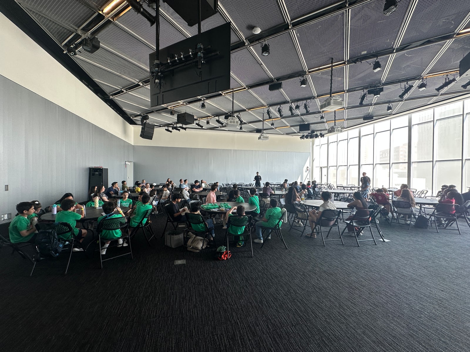 A high-ceilinged room at MIT on Zero Robotics field day, students in green T-shirts at round tables, a wall of windows at the far end