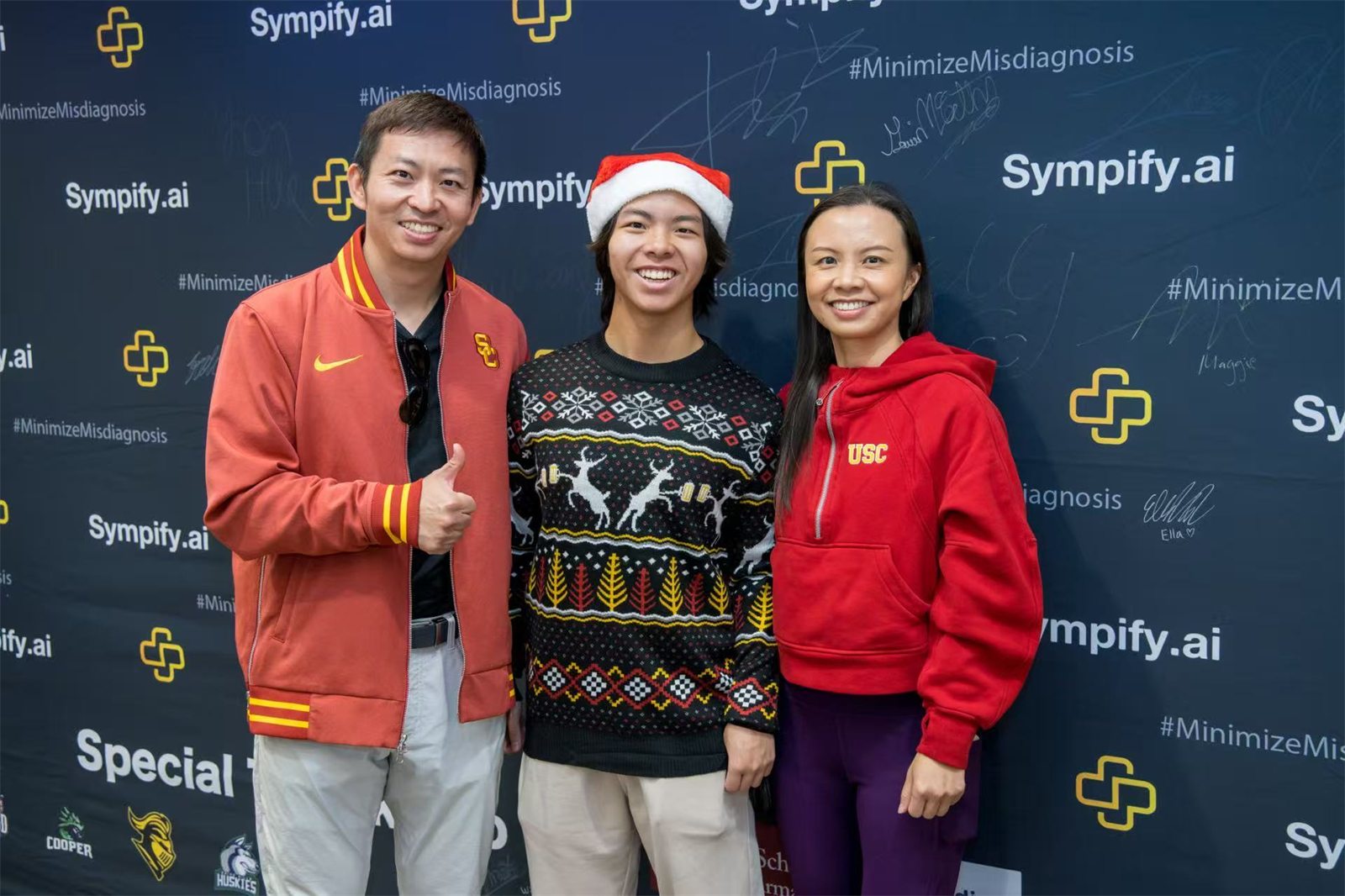 Jalen in a Santa hat and holiday sweater smiling in front of a Sympify.ai step-and-repeat backdrop, flanked by two supporters in USC red