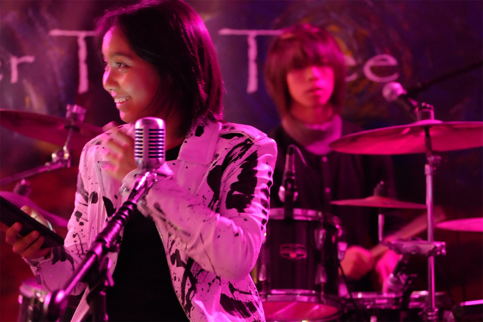 The singer in a paint-splattered white jacket at a vintage microphone, Jalen on drums behind her