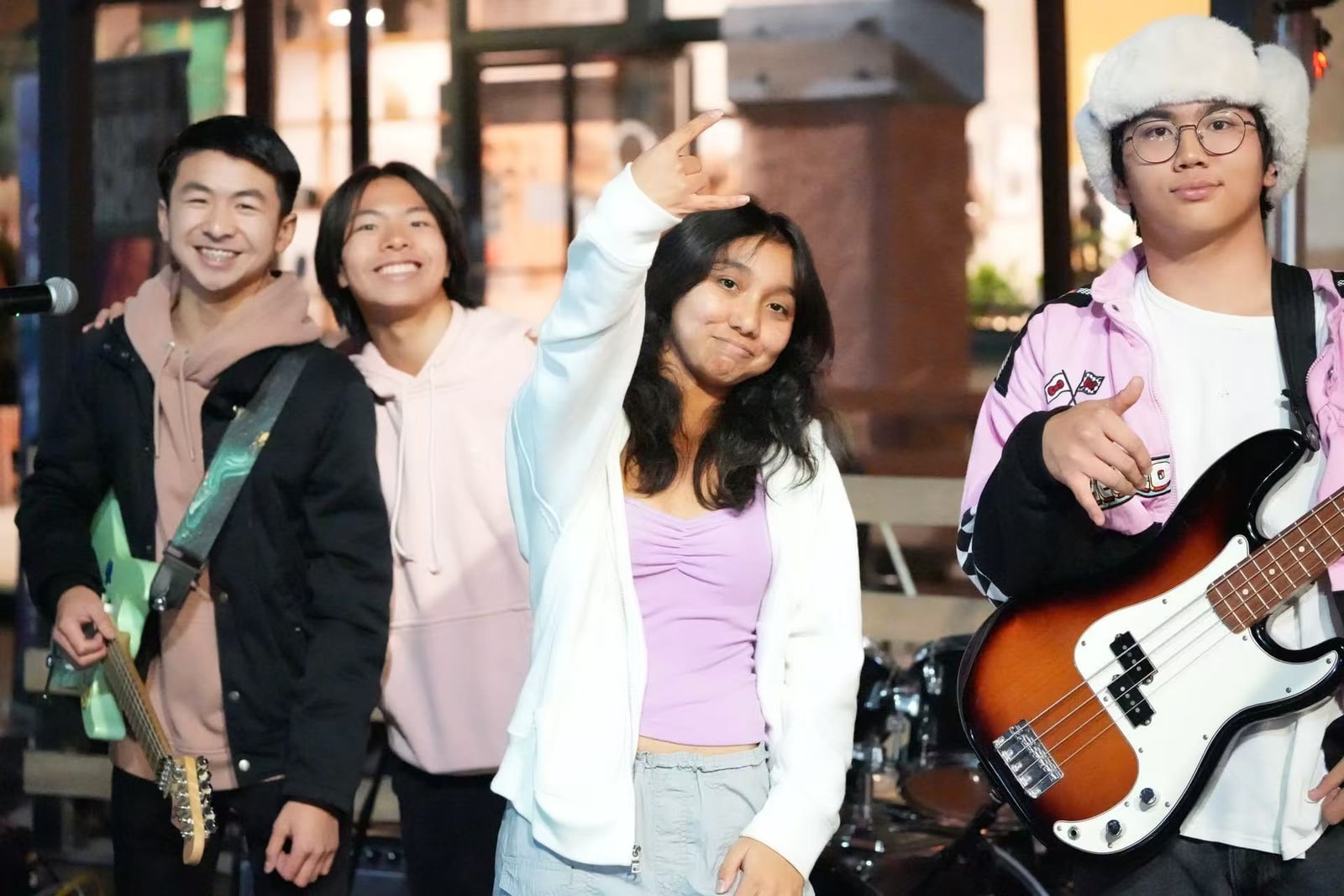 Four members of Post-Credits in front of the night-market stage: two in pink hoodies, one holding a mint guitar, the singer in a white jacket throwing a rock hand, and the bassist in a fluffy white hat