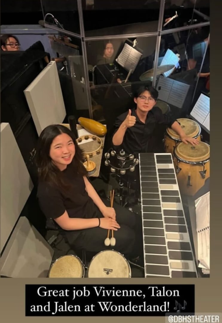 A DBHS Music photo from the Wonderland pit: two percussionists in black among music stands, one at a drum kit with mallets, one giving a thumbs-up behind a marimba and congas, captioned Great job Vivienne, Talon and Jalen at Wonderland