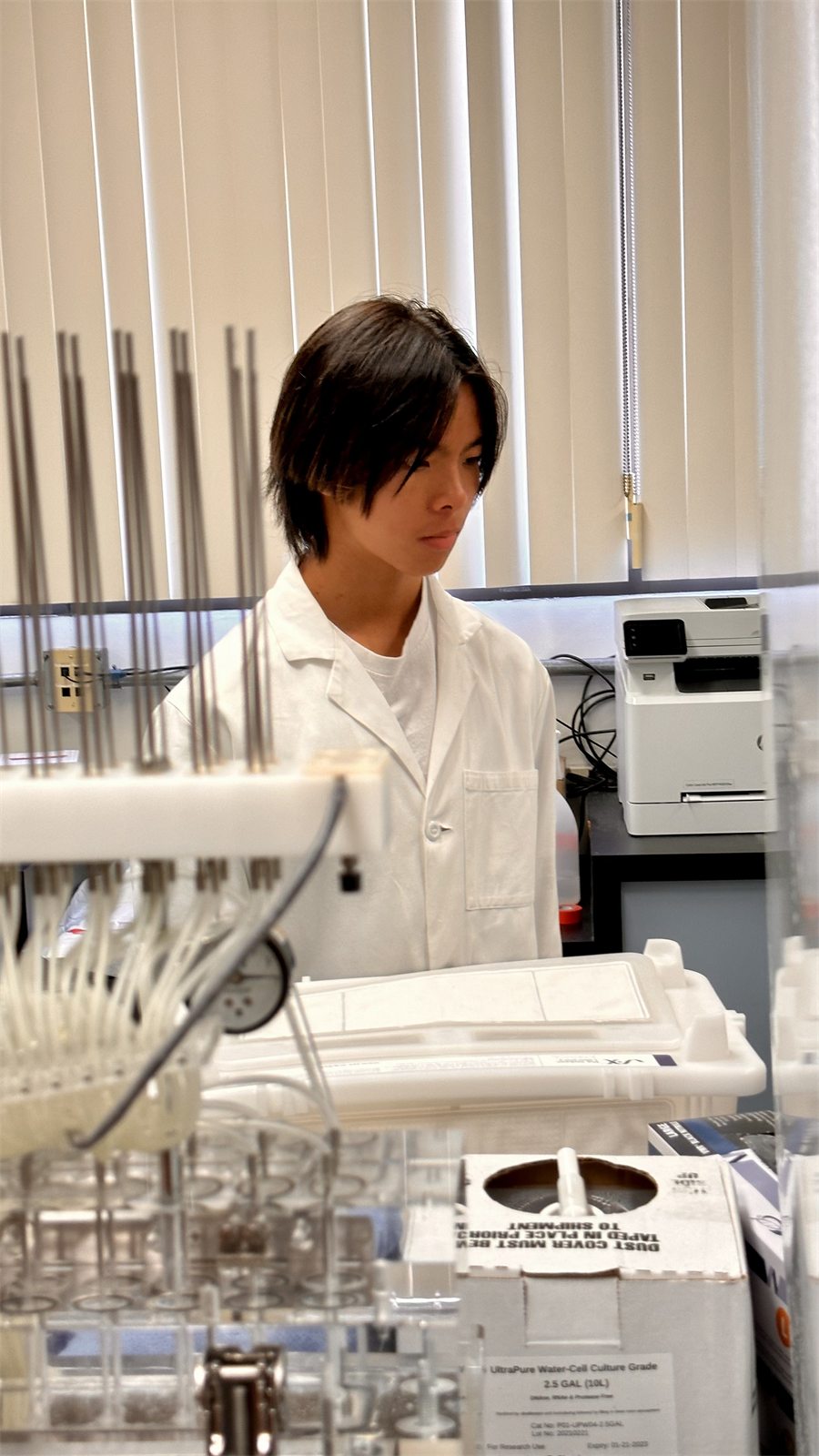 Jalen in a white lab coat at a bench, behind racks of pipette tips and a carboy of cell-culture water