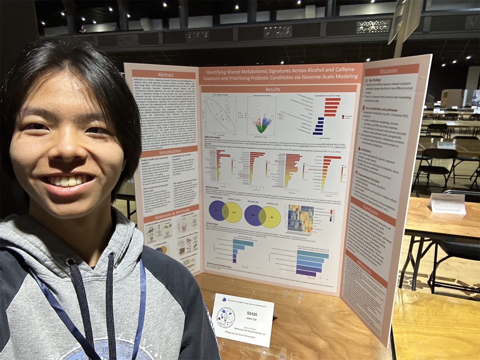 Jalen smiling beside his tri-fold poster, Identifying Shared Metabolomic Signatures Across Alcohol and Caffeine Exposure, in the fair hall