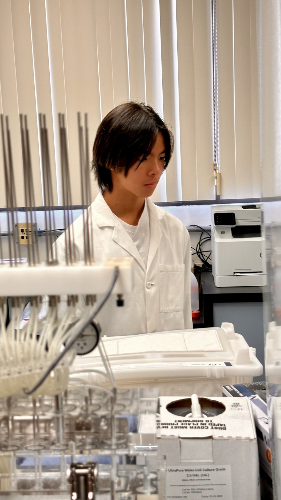 Jalen in a white lab coat looking down at a rack of pipettes, a printer and shelving behind him