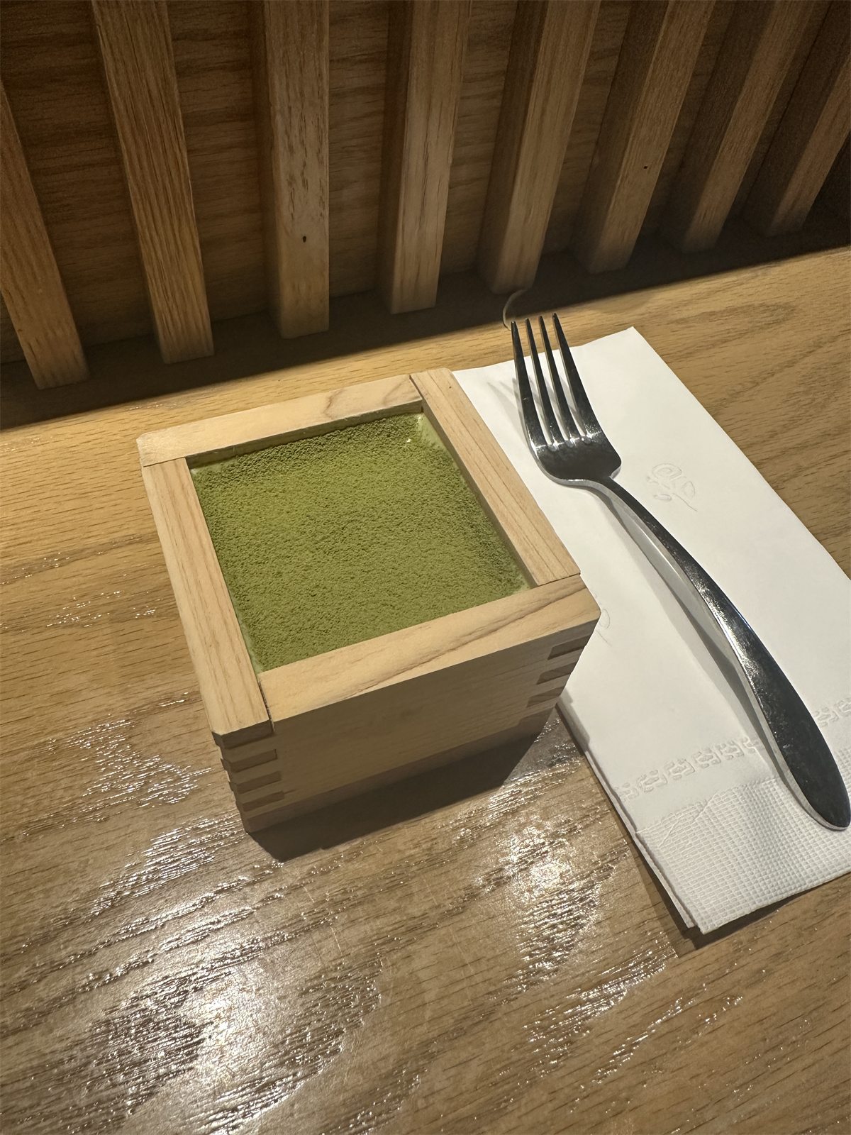 A square wooden masu box filled with matcha tiramisu, a fork resting on a napkin beside it