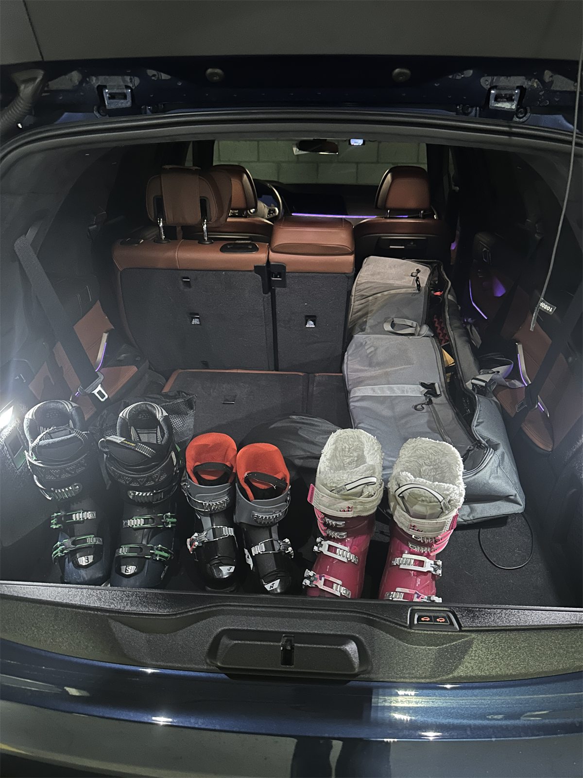 Three pairs of snow boots lined up in the trunk of a car next to a board bag, before a drive to the mountains