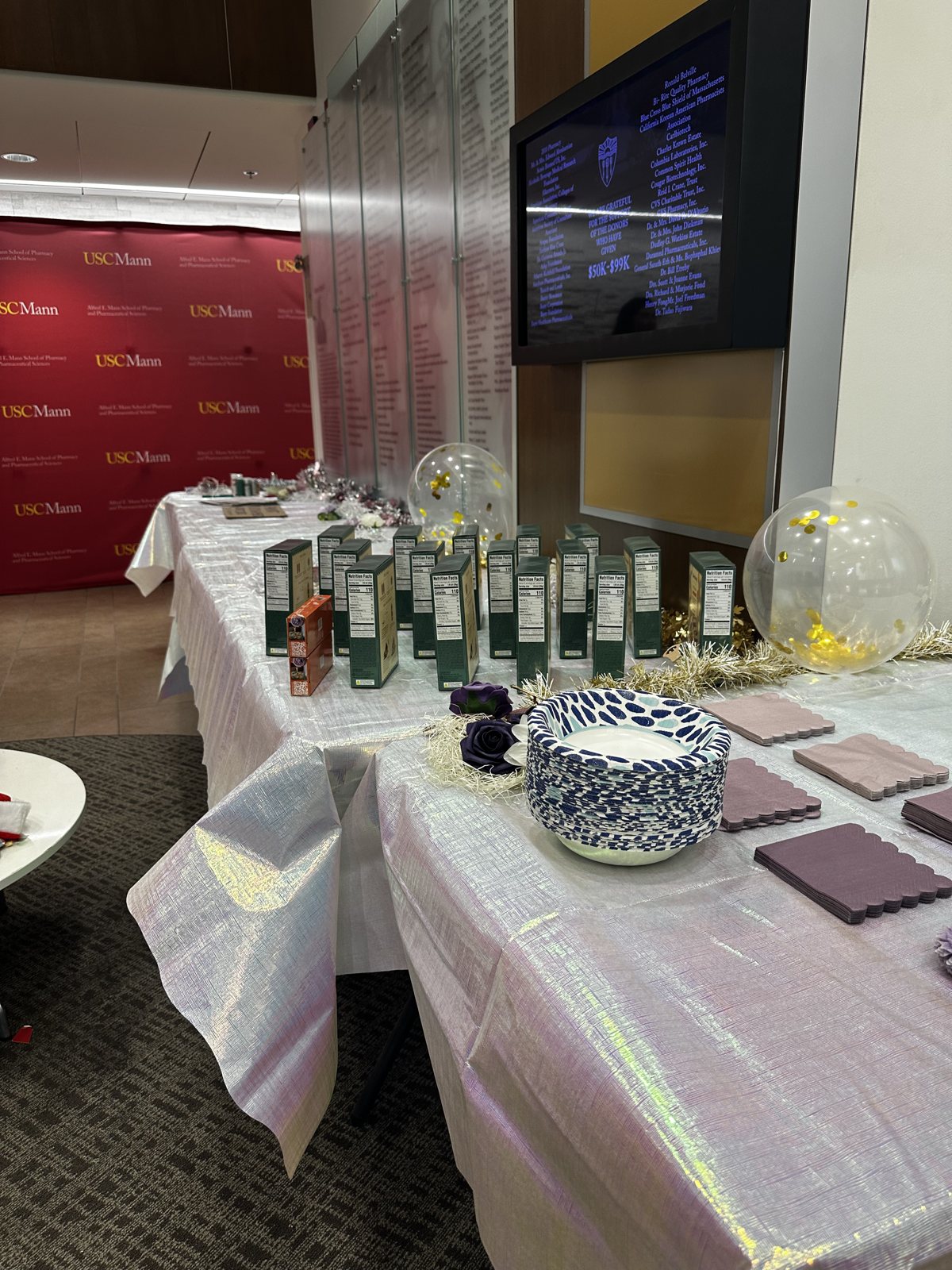 A welcome table dressed in white with rows of green gift boxes and balloons, a USC Mann backdrop behind