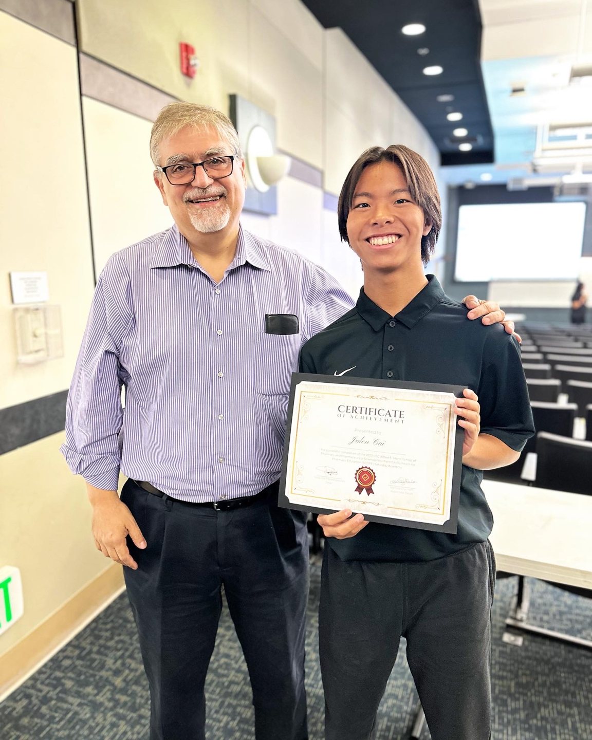 Jalen holding a certificate beside a professor at the front of the lecture hall
