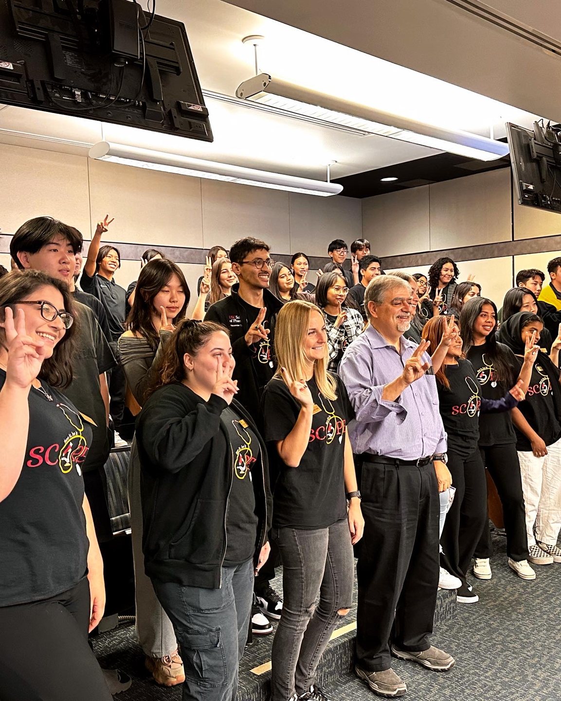 The SCOPE cohort in black T-shirts crowded at the front of the lecture hall with a professor