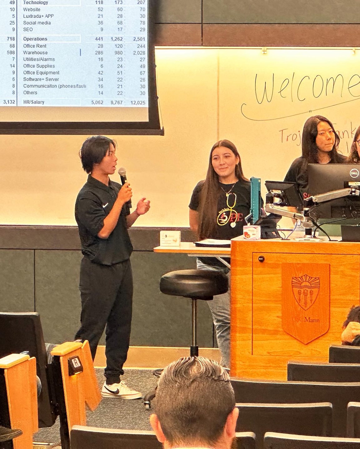 Jalen speaking into a microphone at the front of a lecture hall, Welcome written on the whiteboard behind